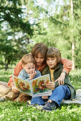 Wall Mural - Vertical portrait of mother hugging two boys while reading book together sitting on picnic blanket in green park