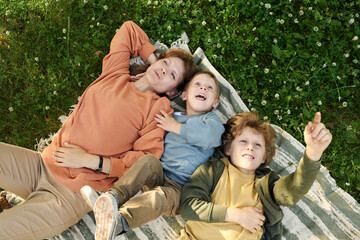 Wall Mural - Top view of mother and two boys lying on picnic blanket together and daydreaming looking at sky and clouds pointing up 