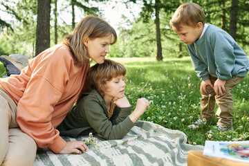 Wall Mural - Candid scene of mother lying on blanket with two young boys in park looking at clover flowers and enjoying nature together
