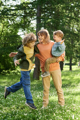 Wall Mural - Vertical full length portrait of young boy jumping up and playing with mother and brother enjoying time together outdoors in green park 