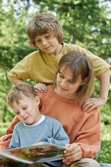 Wall Mural - Vertical portrait of mother with two boys reading book outdoors in sunlit green park with muted color palette