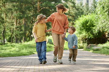 Wall Mural - Full length front view of cheerful mother with two boys holding hands while walking towards camera in green park copy space