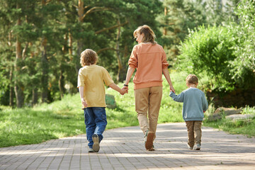 Wall Mural - Full length back view of mother with two boys holding hands and walking away from camera in green park copy space