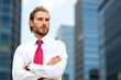 © Minerva Studio - Confident businessman standing with arms crossed in front of skyscrapers