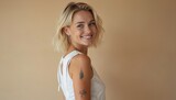 Smiling blond pretty girl with tattoo looking at camera. Beautiful face, healthy skin, short blonde hair. White top. Young female model at beige background. Beauty, skin care, cosmetics, cosmetology,