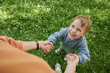 Wall Mural - Candid close-up of smiling young boy looking up at mother while holding hands and playing together in green park outdoors copy space