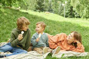 Wall Mural - Portrait of mother with two little boys enjoying time outdoors in park and sitting on picnic blanket with kids eating ice cream