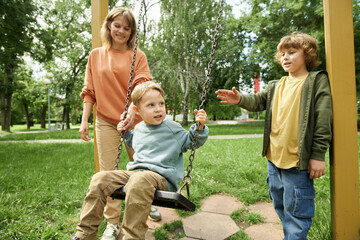 Wall Mural - Portrait of happy family with two children at playground in park, with boy pushing little brother on swing and enjoying outdoor activities together