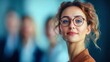 © NovaVision - A confident businesswoman with glasses smiles in a professional setting, surrounded by colleagues.