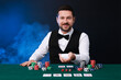 © New Africa - Professional croupier with playing cards at gambling table against color background with smoke