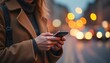 © natakot - A stylish woman in a cozy brown coat is engrossed in her smartphone, surrounded by a vibrant cityscape illuminated by soft, glowing lights. The scene captures the essence of urban life, blending