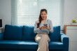 © MINAE - Young Asian businesswoman wearing a gray shirt and beige pants, sitting on a blue sofa in a modern apartment, using a smartphone for work and leisure