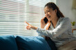 © MINAE - Businesswoman sitting comfortably on a sofa, chatting on her phone while pointing at the window blinds, laughing joyfully and enjoying a lively conversation in a bright, modern apartment