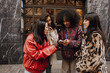 © Drobot Dean - On the street, against the building, a young Black woman checks her phone as three female friends gather around, looking at the screen. Concept of a women's meeting, getting advice from friends.