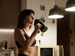 © Drobot Dean - A young, lean Caucasian woman aged 25, wearing a black sports bra, standing in a dimly-lit kitchen at home and drinking some water from a jug, with a confident expression on her face
