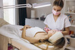 © anatoliycherkas - Beautician wearing mask and gloves injecting filler into lips of young woman lying on bed in cosmetology clinic with magnifying lamp