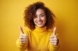 © miss irine - Happy young woman shows thumbs up with smile isolated on yellow background. Joyful lady with curly hair wearing yellow sweater demonstrates approval. Concept of success positive feedback.