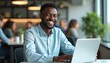 © miss irine - Smiling black man works laptop at desk. Happy businessman wearing blue shirt, looking at camera. Modern office, light interior, professional workspace, remote job.