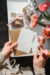 © Elen Nika - Woman holding blank white card above brown envelope arranged above wrapped gifts and pink tulips on white table, suggesting preparation for sending message for special occasion. Greeting card mockup