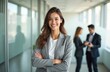 © miss irine - Young business woman smiles confident with arms crossed. Attractive female in formal grey suit stands in modern office environment. Successful career concept. Colleagues communicate at background.