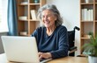 © miss irine - Happy senior woman with grey hair on wheelchair using laptop computer for video call at home. Mature elderly female talking online, smiling, working remotely, telecommuting with modern tech.