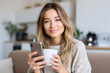 © Chebix - A woman is sitting on a couch holding a cell phone and a cup of coffee