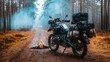© somsri - A fully equipped adventure bike standing near a forest clearing with a smoldering fire.