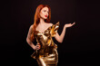 © Beauty Hero - Elegant young woman in a golden dress with red hair and makeup posing against a dark background presenting style