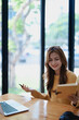 © Jirapong - Smiling young woman engaged in a joyful online e-learning video call, gestu while holding a book with headphones on a wooden table nearby