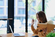 © Jirapong - Smiling young woman engaged in a joyful online e-learning video call, gestu while holding a book with headphones on a wooden table nearby
