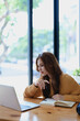 © Jirapong - Smiling young woman engaged in a joyful online e-learning video call, gestu while holding a book with headphones on a wooden table nearby