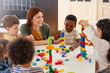 © lordn - Diverse preschool children play with colorful building blocks at a classroom table, guided by a smiling teacher in a creative, inclusive early learning environment.