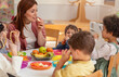 © lordn - A diverse group of preschool children sits around a table eating healthy snacks while a teacher engages with them, promoting nutrition, inclusion, and social interaction in early education.
