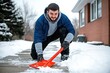 © Wannaeka - Man shoveling snow with red shovel in snowy urban scene, brick house and trees in background on overcast day