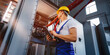 © Olha - Young electrician works with a tablet standing near an industrial fuse board cabinet. Young electrician at work checking equipment inspecting high voltage power electric line distribution fuseboard.