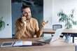 © Liubomir - A smiling businessman is on a phone call, gesturing with his hand. He is seated at a desk with a laptop and other office supplies visible.