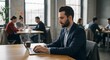 © Matheus - A man in a suit works on a laptop in a bustling, modern office space, with colleagues collaborating at desks in the background.