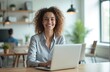 © miss irine - Joyful businesswoman working on laptop in modern office. Smiling young woman with curly hair in casual wear looking at camera. Positive cheerful workplace atmosphere with copy space.