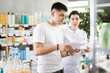 © JackF - Young guy with medical prescription in hand looking for something in big drugstore, young girl pharmacist consulting and helping client