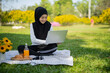 © Wanwajee - Young Asian Muslim female student sitting on mat, using notebook at summer garden park. Isalamic business woman enjoy relaxing with laptop and picnic at public.