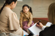 © DragonImages - Two women discussing childs progress in kindergarten education with child present in blurred background, adult taking notes during conversation with family