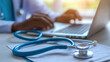 © Ivan - A health care worker is focused on a laptop, likely processing patient information or conducting research. A stethoscope rests nearby on documents. The setting is bright and professional