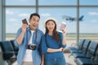 © Mallika - Happy young Asian couple holding passports and boarding passes at the airport terminal, excited and ready for their trip. Travel and vacation concept with background of airplane taking off.