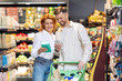 © Home-stock - Spouses doing grocery shopping in supermarket on weekend, using cellphone for checking sales, standing in store aisles indoors