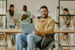 © AnnaStills - Portrait of handsome young IT specialist with disability holding laptop of knees smiling at camera while his colleagues working in background