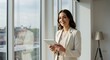 © alleb - Woman in business attire holds a tablet standing near a large window.
