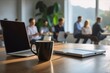 © YiuCheung - Laptop, coffee mug, and notebook on a wooden desk in a modern office.  Sunlight streams through large windows, illuminating the scene. Blurred colleagues in the background are engaged in a meeting or