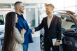 © Anastasiya - Side view of smiling couple shaking hands with salesman, happy young family buying new car, luxury vehicles displayed inside the spacious and brightly lit dealership showroom, closeup