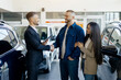 © Anastasiya - Friendly salesman shaking hands with a smiling couple inside a bright, modern car dealership center, cars visible around them. Happy young family buying new vehicle in auto salon