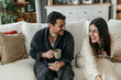 © La Famiglia - Happy couple wearing bathrobes, using smartphones, drinking coffee and laughing on the sofa at home, enjoying a relaxing weekend morning